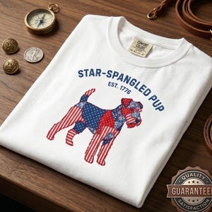 May include: A white t-shirt featuring a graphic of a dog with a red, white, and blue patriotic pattern. The text above the dog reads STAR-SPANGLED PUP EST. 1776 in blue letters. The shirt has a Comfort Colors brand label on the inside neck area.
