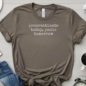 May include: A brown t-shirt with the text "procrastinate today, panic tomorrow" in white. The shirt is knotted at the bottom. Other items include a pair of blue jeans, a smartphone, a cup of coffee, and a black alarm clock.