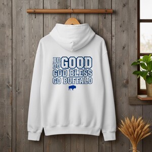Puede incluir: Sudadera con capucha blanca con el texto "BE GOOD GOD BLESS GO BUFFALO" en azul. Una silueta de b&uacute;falo azul est&aacute; debajo del texto. La sudadera con capucha est&aacute; colgada en una percha de madera sobre un fondo de madera.