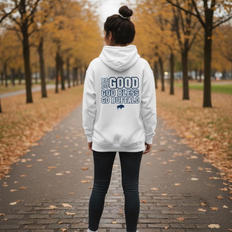 Puede incluir: Una sudadera con capucha blanca con el texto "BE DO GOOD GOD BLESS GO BUFFALO" en azul. La sudadera presenta un peque&ntilde;o logotipo de b&uacute;falo azul. La persona lleva pantalones negros y est&aacute; de pie en un camino bordeado de &aacute;rboles.