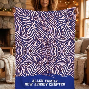 May include: A large, rectangular blanket with a white and navy blue abstract pattern. The blanket has a blue border with the text "ALLEN FAMILY NEW JERSEY CHAPTER" in white. The blanket is held up by a person.