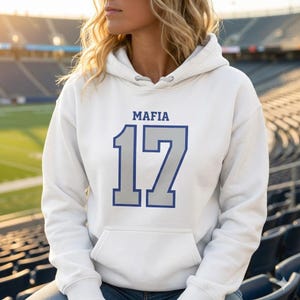 Puede incluir: Sudadera con capucha blanca con la palabra "MAFIA" encima del n&uacute;mero "17" en azul y gris. La sudadera tiene un bolsillo delantero y una capucha con cord&oacute;n. El fondo muestra un estadio.