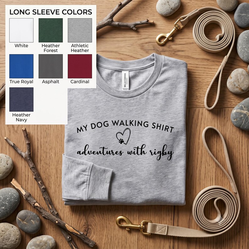 K&ouml;nnte beinhalten: Ein hellgraues Langarmshirt mit schwarzem Text, der MY DOG WALKING SHIRT lautet, einem Herzsymbol mit Pfotenabdruck und dem Text adventures with rigby. Eine Farbkarte zeigt die Optionen Wei&szlig;, Heather Forest, Athletic Heather, True Royal, Asphalt, Cardinal und Heather Navy.