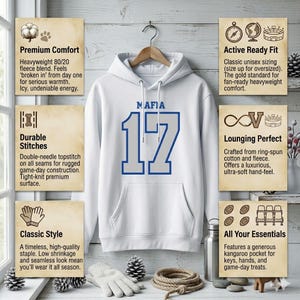 Puede incluir: Sudadera con capucha blanca con la palabra "MAFIA" encima del n&uacute;mero "17" en azul. La sudadera tiene un bolsillo de canguro y una capucha con cord&oacute;n. La sudadera est&aacute; colgada de una percha de madera.