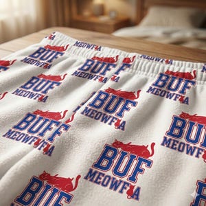 May include: White fleece pajama bottoms featuring a repeating pattern of red cats and the text "BUF MEOWFA" in blue. The trousers have a drawstring waist and are made of a soft, textured fabric.