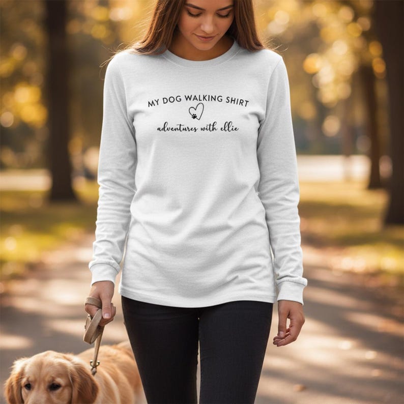 K&ouml;nnte beinhalten: Ein wei&szlig;es Langarmshirt mit schwarzem Text, der MY DOG WALKING SHIRT lautet, einem Herzsymbol mit Pfotenabdruck und dem Text adventures with ellie. Das Shirt wird mit einer schwarzen Hose kombiniert.