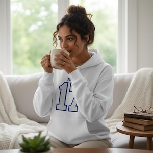 Puede incluir: Sudadera con capucha blanca con el n&uacute;mero 14 azul en la parte delantera. La sudadera con capucha tiene una capucha con cord&oacute;n y mangas largas. La persona sostiene una taza blanca.