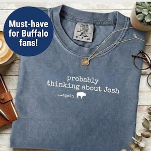 May include: A blue-grey t-shirt with the text "probably thinking about Josh...again" and a buffalo graphic. A gold necklace and glasses are on the shirt. The shirt has a tag that says "Comfort Colors".