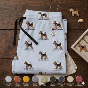May include: Light blue pajama pants with a black drawstring, featuring a repeating pattern of a dog with a duck on its back, and the text "we ride at dawn". Gold-colored jewelry and dog pins are also visible.