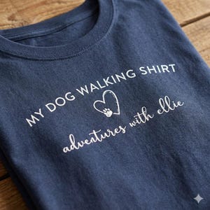 K&ouml;nnte beinhalten: Ein marineblaues T-Shirt mit wei&szlig;em Siebdrucktext, der MY DOG WALKING SHIRT lautet, einem Herzumriss mit einer Pfotenabdruck-Grafik darin und dem Text adventures with ellie in einer kursiven Schriftart auf der Brust.