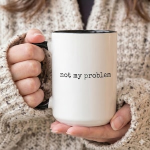May include: A white ceramic mug with a black interior and handle. The exterior features the text "not my problem" printed in a black, typewriter-style font centred on the front of the mug. The mug has a cylindrical shape and a smooth finish.
