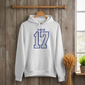 Puede incluir: Una sudadera con capucha blanca con la palabra "MAFIA" encima del n&uacute;mero "17" en azul. La sudadera tiene un bolsillo delantero y una capucha con cord&oacute;n. El dise&ntilde;o est&aacute; centrado en la parte delantera de la prenda.