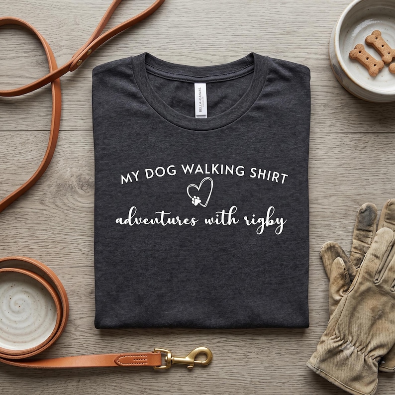 K&ouml;nnte beinhalten: Ein dunkel anthrazitfarbenes T-Shirt mit wei&szlig;em Text, der MY DOG WALKING SHIRT lautet, sowie einem Herz- und Pfotenabdruck-Symbol &uuml;ber den in Schreibschrift gehaltenen W&ouml;rtern adventures with rigby, die mittig auf der Vorderseite stehen.