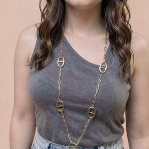 Puede incluir: Un collar largo de color dorado con eslabones grandes de forma ovalada y un diseño de cadena. El collar se lleva sobre una camiseta sin mangas gris y unos vaqueros azules. El collar tiene un colgante ovalado grande en la parte inferior.