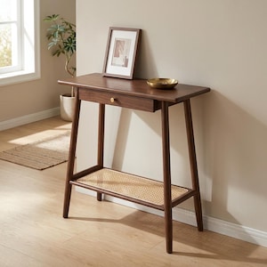 Può includere: Un tavolino consolle in legno marrone scuro con un cassetto e un ripiano inferiore con un inserto in rattan intrecciato. Un'immagine incorniciata e una piccola ciotola dorata sono in cima. Il tavolo ha quattro gambe sottili e angolate e un piccolo pomello dorato sul cassetto.