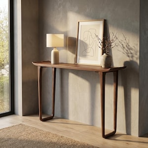 May include: A dark brown wooden console table with a curved top and legs. A beige lamp, framed artwork, and a vase with branches sit on the table. The room has a neutral color palette and natural light.