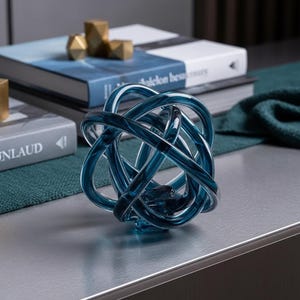 Puede incluir: Una escultura decorativa de nudo de vidrio azul. La escultura está hecha de varillas de vidrio curvas e entrelazadas, creando un diseño complejo e interconectado. La escultura está sobre una superficie plateada, con libros y objetos geométricos dorados en el fondo.