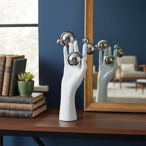May include: A white ceramic hand sculpture holding several shiny silver spheres of varying sizes. The sculpture is displayed on a dark brown surface, with a mirror reflecting the hand and spheres. A small succulent in a green pot and stacked books are visible.