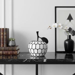 Può includere: Una scultura decorativa a forma di mela con corpo bianco e motivo geometrico nero. La mela ha un gambo e una foglia neri. È esposta su una superficie di marmo nero, con libri e un quadro incorniciato sullo sfondo.