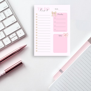 Könnte beinhalten: Ein rosa-weißes To-Do-Listen-Notizblock mit einer Schleifengrafik, einer Tastatur und zwei Stiften. Der Notizblock hat Abschnitte für Datum, Prioritäten und Notizen. Die Stifte und die Tastatur sind roségoldfarben.