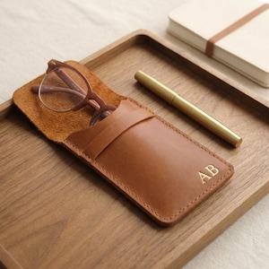 Könnte beinhalten: Eine braune Leder-Brillenetui mit den goldenen Initialen "AB". Darin befindet sich eine Brille mit braunem Rahmen. Ein goldener Stift und ein Notizbuch liegen auf einem Holztablett.