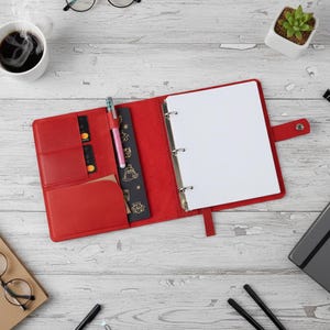 Op de afbeelding: Open rode leren binder met een drievoudig ringmechanisme, met een pen, kaartsleuven en een bladwijzer. De binder is open om blanco wit papier te onthullen. Een kopje koffie en een kleine plant zijn ook zichtbaar.
