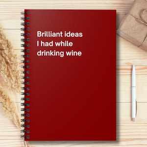 May include: A red spiral-bound notebook with the text "Brilliant ideas I had while drinking wine" in white. A wrapped gift, a white pen, and dried wheat stalks are also on the wooden surface.