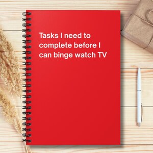 May include: A red spiral-bound notebook with the text "Tasks I need to complete before I can binge watch TV." A pen, a wrapped gift, and dried plants are also on the wooden surface.
