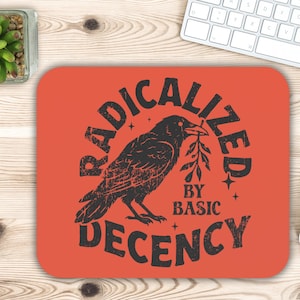 May include: A rectangular mousepad with rounded corners in a vibrant orange hue. The design features a black raven holding a sprig, surrounded by the words "RADICALIZED BY BASIC DECENCY" in a distressed font.
