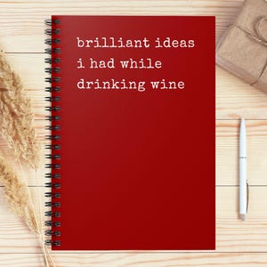 May include: A red spiral-bound notebook with the text "brilliant ideas i had while drinking wine" in white. A white pen, a wrapped gift, and dried wheat stalks are also on the light wood surface.