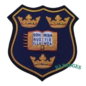May include: A navy blue embroidered shield-shaped patch with three gold crowns and an open book bearing the Latin inscription "DOMI MINA NVS TIO ILLU MEA". The patch is edged with gold thread and has the text "RA BADGES" in the bottom right corner.