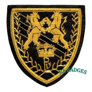 Puede incluir: Parche bordado en negro y dorado con forma de escudo. Presenta dos leones dorados enfrentados, una corona dorada y las letras "R" y "B". El borde y los detalles son dorados. El texto "RABBADGES" es visible.