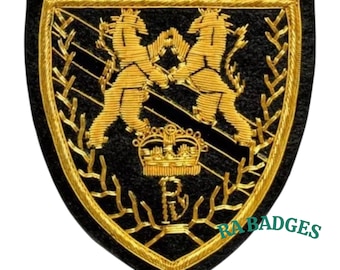 May include: Black and gold embroidered patch with a shield shape. The patch features two gold lions facing each other, a gold crown, and the letters "R" and "B". The border and details are gold.