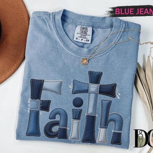 A blue denim t-shirt with the word "Faith" printed on it, along with a cross and other religious symbols, is displayed on a white surface.