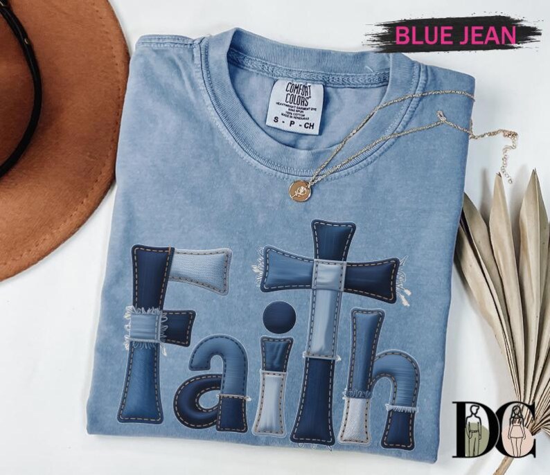 A blue denim t-shirt with the word "Faith" printed on it, along with a cross and other religious symbols, is displayed on a white surface.
