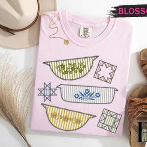 May include: A light pink t-shirt with embroidered casserole dish designs in green, blue, and yellow. The shirt has a crew neck and the word "Blossom" in pink. Sandals and a hat are also in the image.
