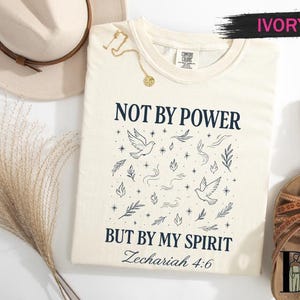 May include: Ivory-colored t-shirt with the text "NOT BY POWER BUT BY MY SPIRIT Zechariah 4:6" in navy blue. The design includes doves, leaves, and stars. A gold necklace and brown sandals are also visible.