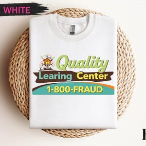 May include: White crew neck t-shirt with the text "Quality Learning Center 1-800-FRAUD" in green, brown, and blue. The design includes a small house illustration. The shirt is displayed on a woven, circular surface.