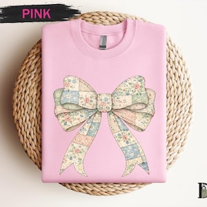 May include: A light pink sweatshirt featuring a large bow design. The bow is a patchwork of floral and patterned fabrics in pastel colors. The word "PINK" is written in black above the sweatshirt.