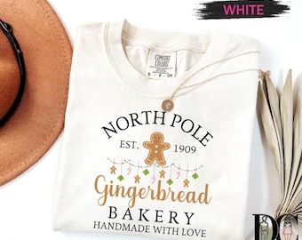 May include: A white Comfort Colors t-shirt with a "North Pole Gingerbread Bakery" design. The design includes a gingerbread man, candy canes, and stars. The text "Handmade with Love" is also included.