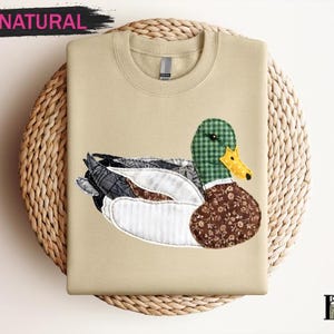 May include: A beige sweatshirt featuring a detailed appliqué of a duck. The duck has a green plaid head, a yellow beak, and a brown floral patterned body. The word "NATURAL" is above the sweatshirt in a black banner.