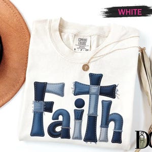 a white t-shirt with a blue cross design and the word "Faith" written on it. The t-shirt is placed on a white background, along with a brown hat and a fan.