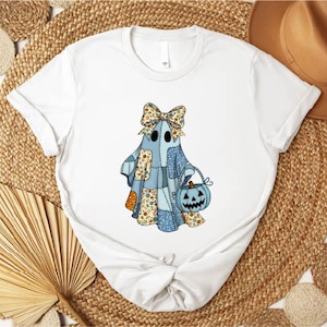May include: White t-shirt featuring a cartoon ghost with a patchwork design in blue, orange, and beige. The ghost wears a bow and carries a blue pumpkin bucket. The shirt is styled on a woven background.