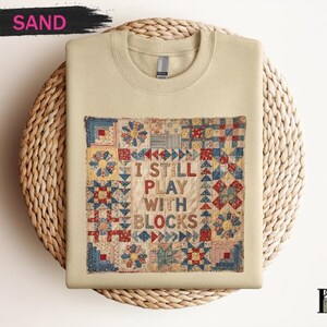May include: Sand-colored sweatshirt with a colorful quilt design. The quilt features red, blue, and yellow patterns, with the text "I STILL PLAY WITH BLOCKS" in the center. The sweatshirt is displayed on a woven mat.