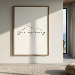 Op de afbeelding: Een ingelijste kunstprint met een minimalistische bergcontour en de tekst "Gone exploring" in handschriftstijl. Het kunstwerk staat tegen een witte achtergrond, is omlijst in een lichte houttint en wordt in een modern interieur getoond.