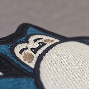 Peut inclure: Un &eacute;cusson brod&eacute; repr&eacute;sentant un personnage stylis&eacute; avec un grand ventre cr&egrave;me, une fourrure bleu fonc&eacute; et des yeux ferm&eacute;s. Le motif est soulign&eacute; par un fil noir &eacute;pais, mettant en valeur des motifs de couture d&eacute;taill&eacute;s sur les zones bleues et cr&egrave;me.