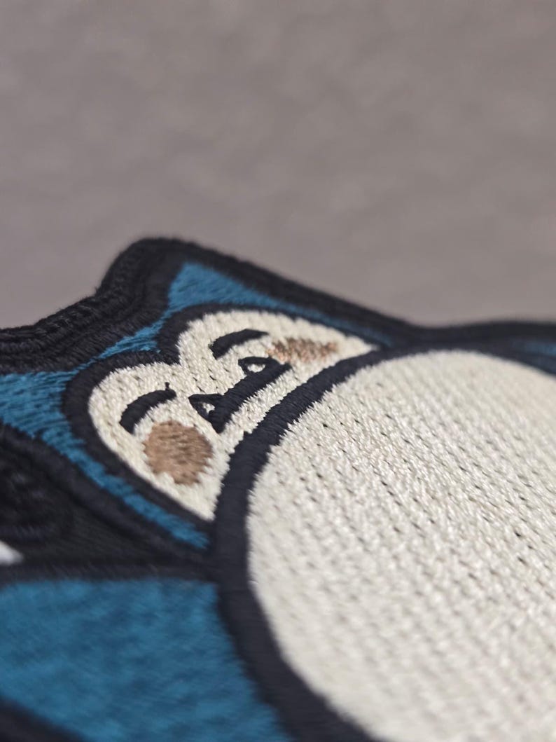 Peut inclure: Un &eacute;cusson brod&eacute; repr&eacute;sentant un personnage stylis&eacute; avec un grand ventre cr&egrave;me, une fourrure bleu fonc&eacute; et des yeux ferm&eacute;s. Le motif est soulign&eacute; par un fil noir &eacute;pais, mettant en valeur des motifs de couture d&eacute;taill&eacute;s sur les zones bleues et cr&egrave;me.