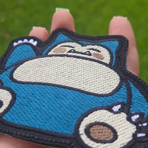 Peut inclure: Un &eacute;cusson brod&eacute; repr&eacute;sentant une cr&eacute;ature bleue et cr&egrave;me avec un gros ventre, les yeux ferm&eacute;s et de petites griffes. Le motif est soulign&eacute; par un fil noir, mettant en valeur des coutures d&eacute;taill&eacute;es sur tout le corps et le visage.