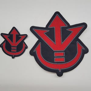 May include: Two embroidered patches featuring a red and black design. The larger patch has a stylized arrow and anchor shape, while the smaller patch mirrors the design. The patches are likely for clothing or accessories.