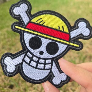 May include: Embroidered patch featuring a skull and crossbones design with a straw hat. The patch has a yellow hat with a red band, black eyes, and white bones. The patch is outlined in black.